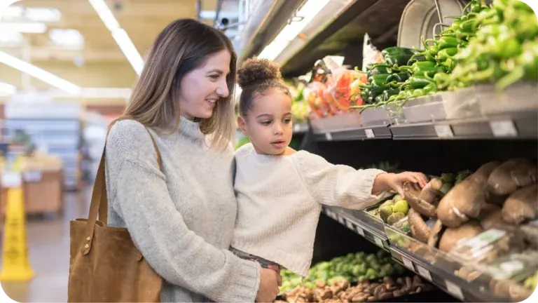 A mother holding her small daughter at a supermarket’s fresh produce shelves, the child reaching out for a lime.