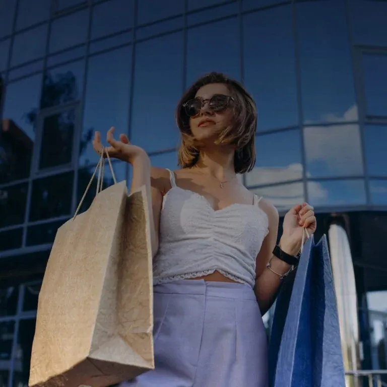 A shopper in sunglasses carrying paper and fabric bags, photographed from below against the glass façade of a shopping centre at dusk.