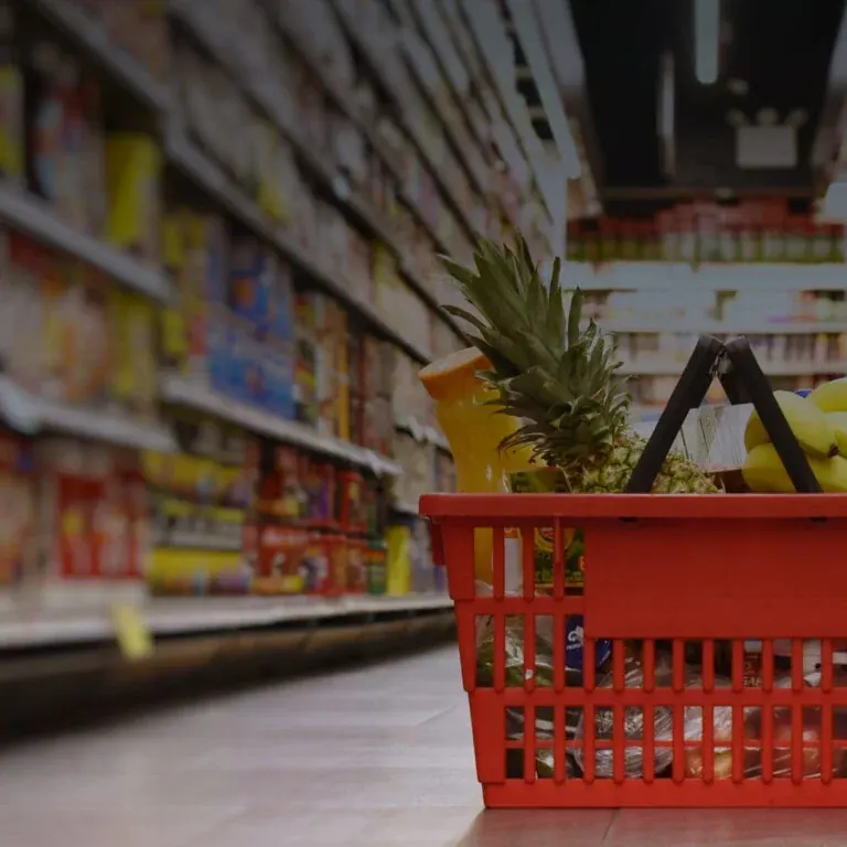A red shopping basket holding a pineapple, bananas and a bottle of juice, photographed low down a supermarket aisle with the shelves blurred behind it.