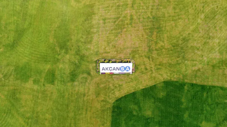 Mown lawns seen straight down from the air, with the AKÇANSA wordmark laid out in a rectangular flowerbed at the centre of them.