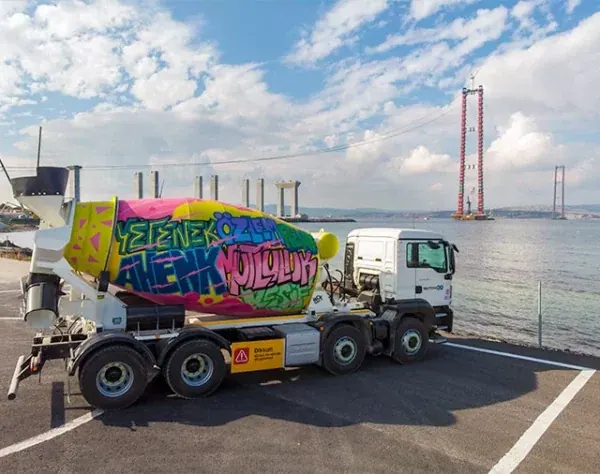 An AkçanSA concrete mixer truck parked on the quayside, its drum hand-painted in bright street-art lettering, with the 1915 Çanakkale bridge under construction behind it.