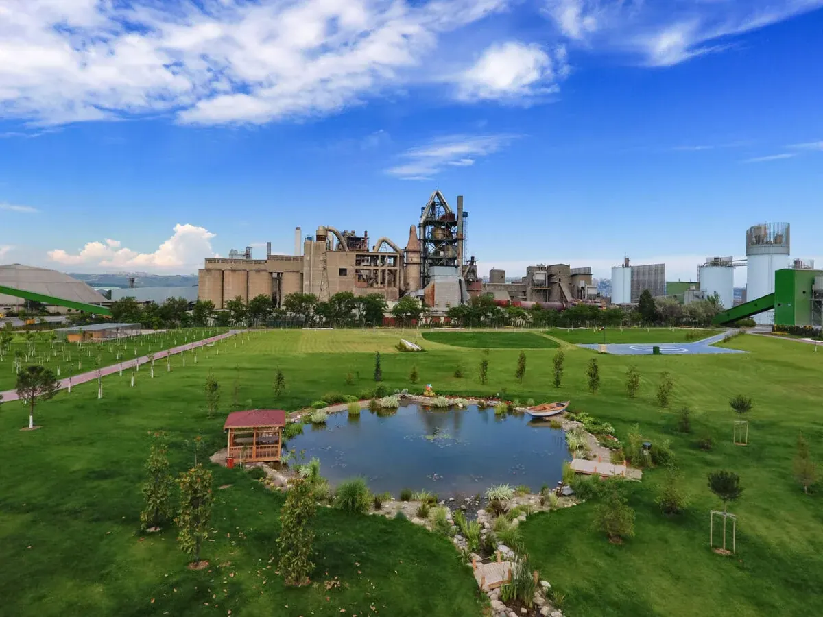 An AkçanSA cement works behind a landscaped park of lawns, young trees and a pond with a wooden gazebo beside it, under a bright blue sky.