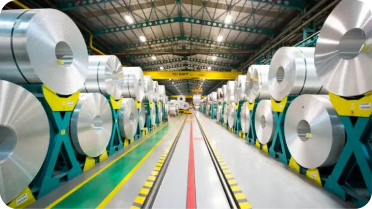 Rows of aluminium coils on teal cradles down both sides of a lit mill aisle, green and red floor lanes running to the far end of the hall.