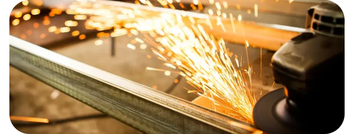 An angle grinder throwing a spray of orange sparks along a steel profile on a workshop bench.