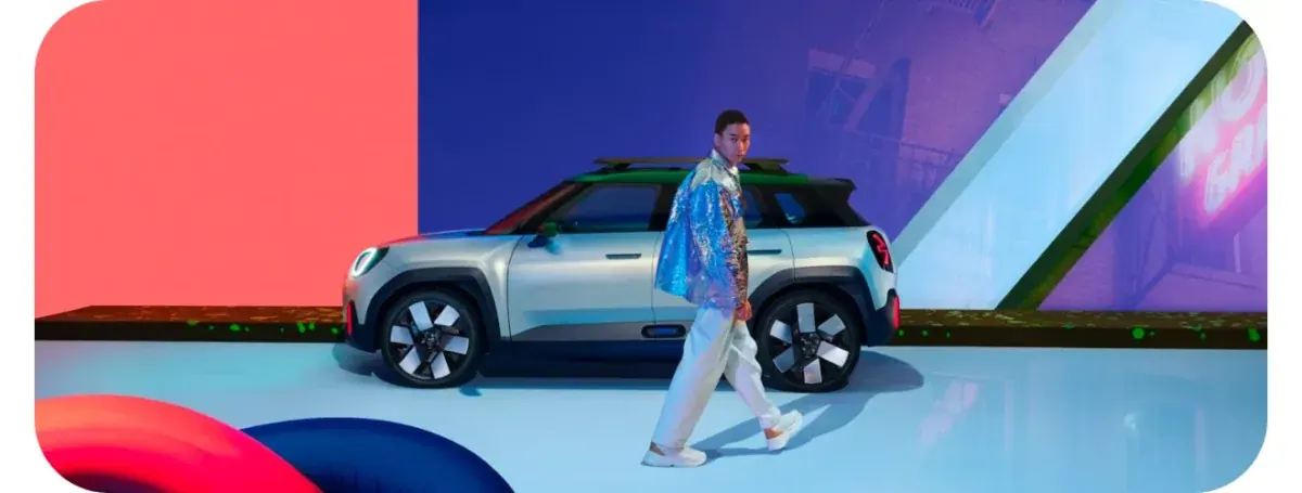 A silver electric MINI with a green roof on a lit studio set of coral, blue and purple panels, a model in a sequinned cape walking past it.