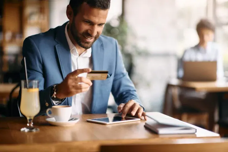 A smiling businessman at a café table holding a bank card in one hand and using a tablet with the other.