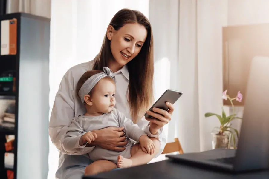 A mother at her desk holding her baby on her lap, reading something on her phone, a laptop open beside her.
