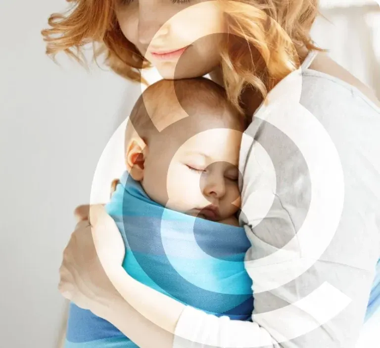 A mother holding her sleeping baby, wrapped in a blue sling against her shoulder.