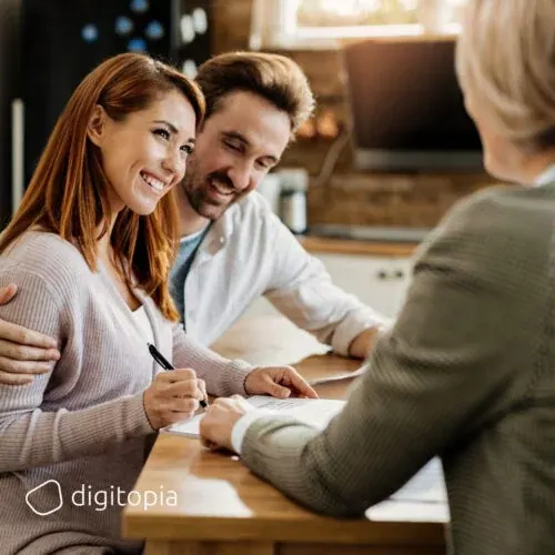 A smiling couple signing paperwork at a kitchen table with their adviser