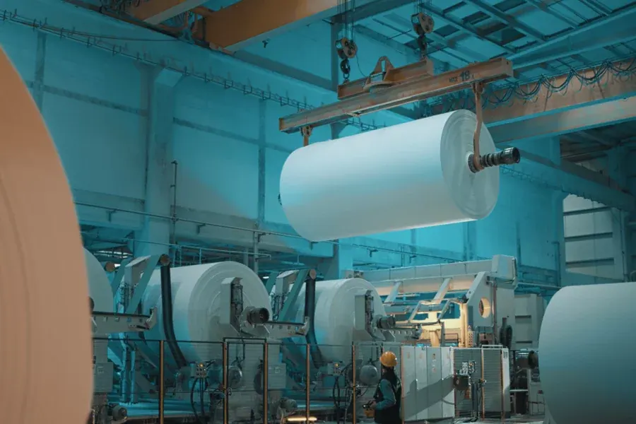 A finished reel of tissue paper hanging from an overhead crane in Lila Kağıt’s mill, above a line of paper machines with a worker in a hard hat beside them.