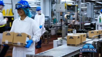 A Litehouse Foods packing line: an Employee Owner in a white coat, hairnet, safety glasses and blue hard hat carrying a stack of cartons past the conveyor.
