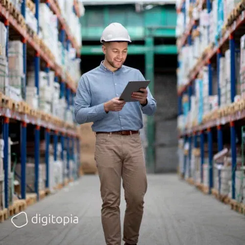 A warehouse worker in a hard hat walking a racking aisle, reading a tablet