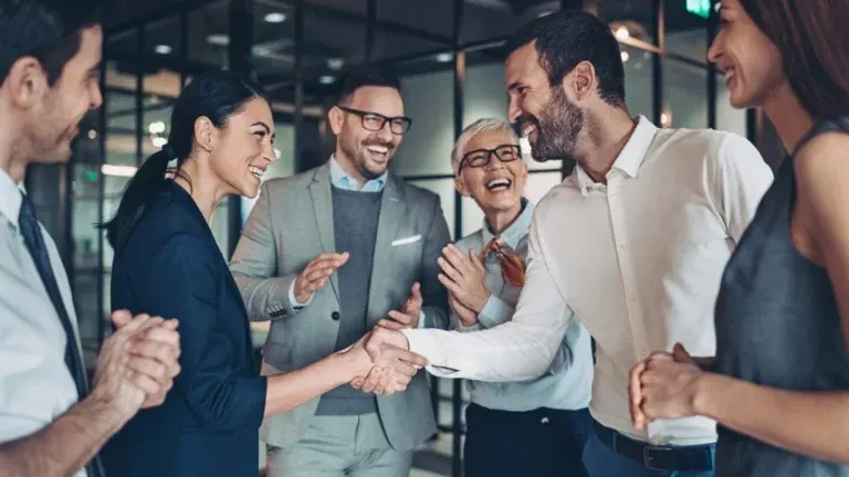 Two consultants shaking hands while their colleagues applaud in a glass-walled office.