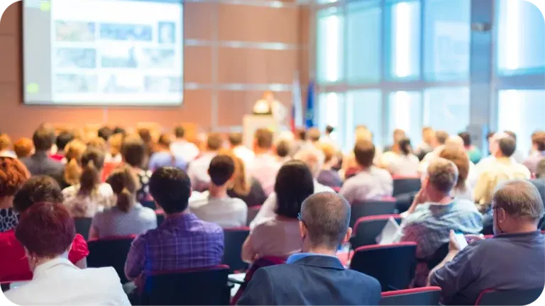 A full congress hall seen from the back: rows of delegates on red chairs facing a speaker at the lectern and a lit presentation screen.