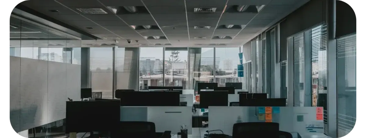 An empty open-plan office at dusk: desks and monitors between glass partitions, with the city visible through the window wall behind them.