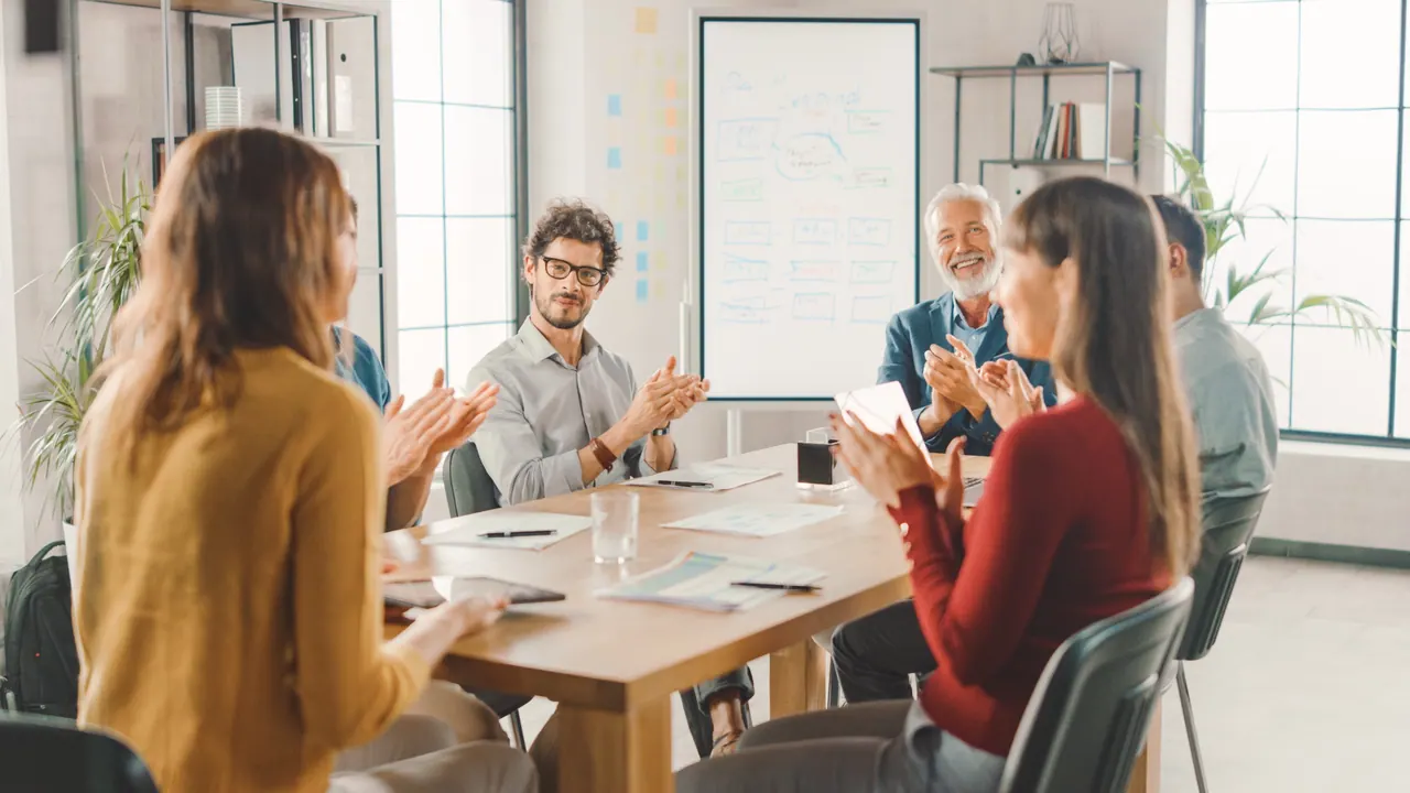 A team applauding around a meeting table at the end of a review, a plan worked out on the whiteboard behind them.