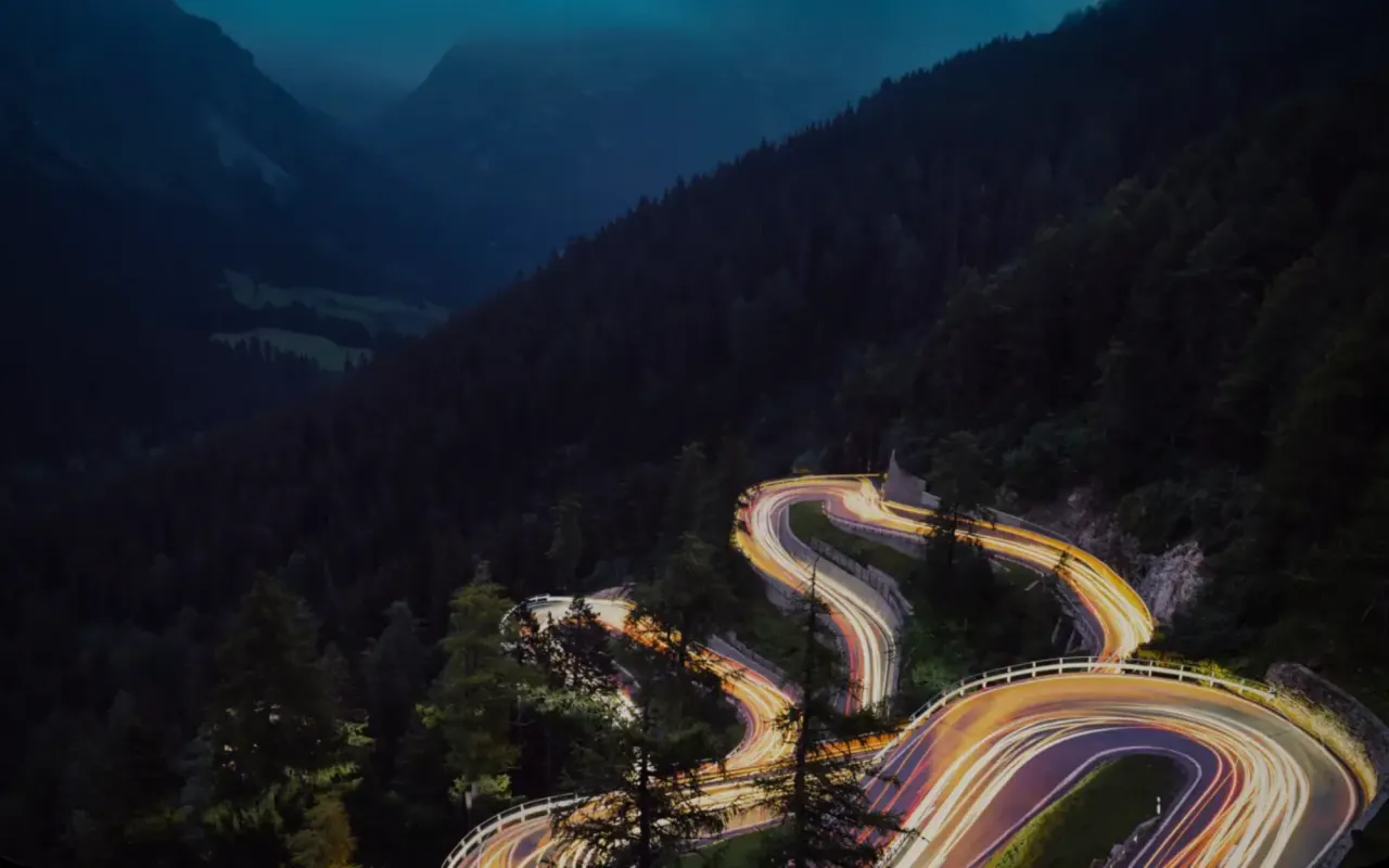 A hairpin mountain road at dusk, the headlights of passing cars drawn into long ribbons of light through the forest.
