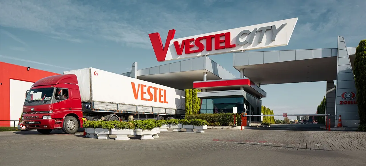 The entrance to Vestel City under a pale sky, the VESTEL CITY sign above the gatehouse and a red Vestel lorry pulling away from it.