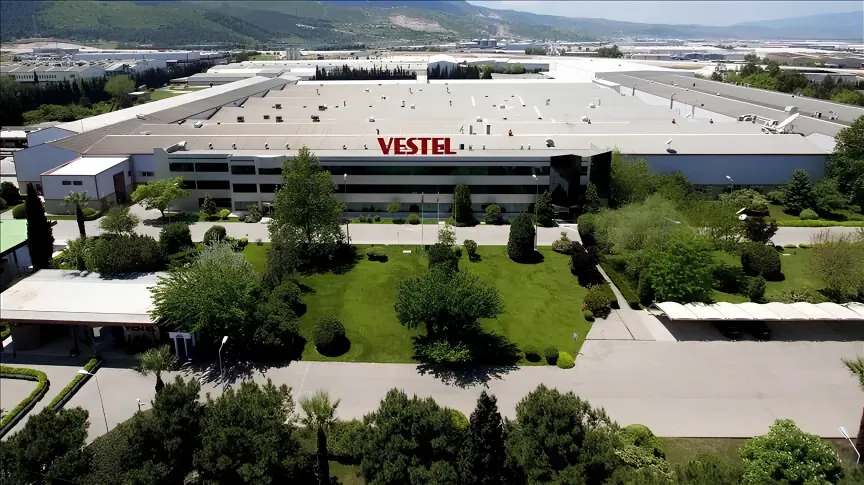 An aerial view of a Vestel plant, its long white roofline carrying the Vestel name, with landscaped grounds in front and hills behind.