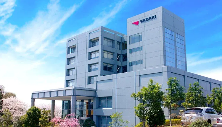 Yazaki’s headquarters: a grey office building carrying the Yazaki mark, under a blue sky, with cherry blossom in the garden in front of it.